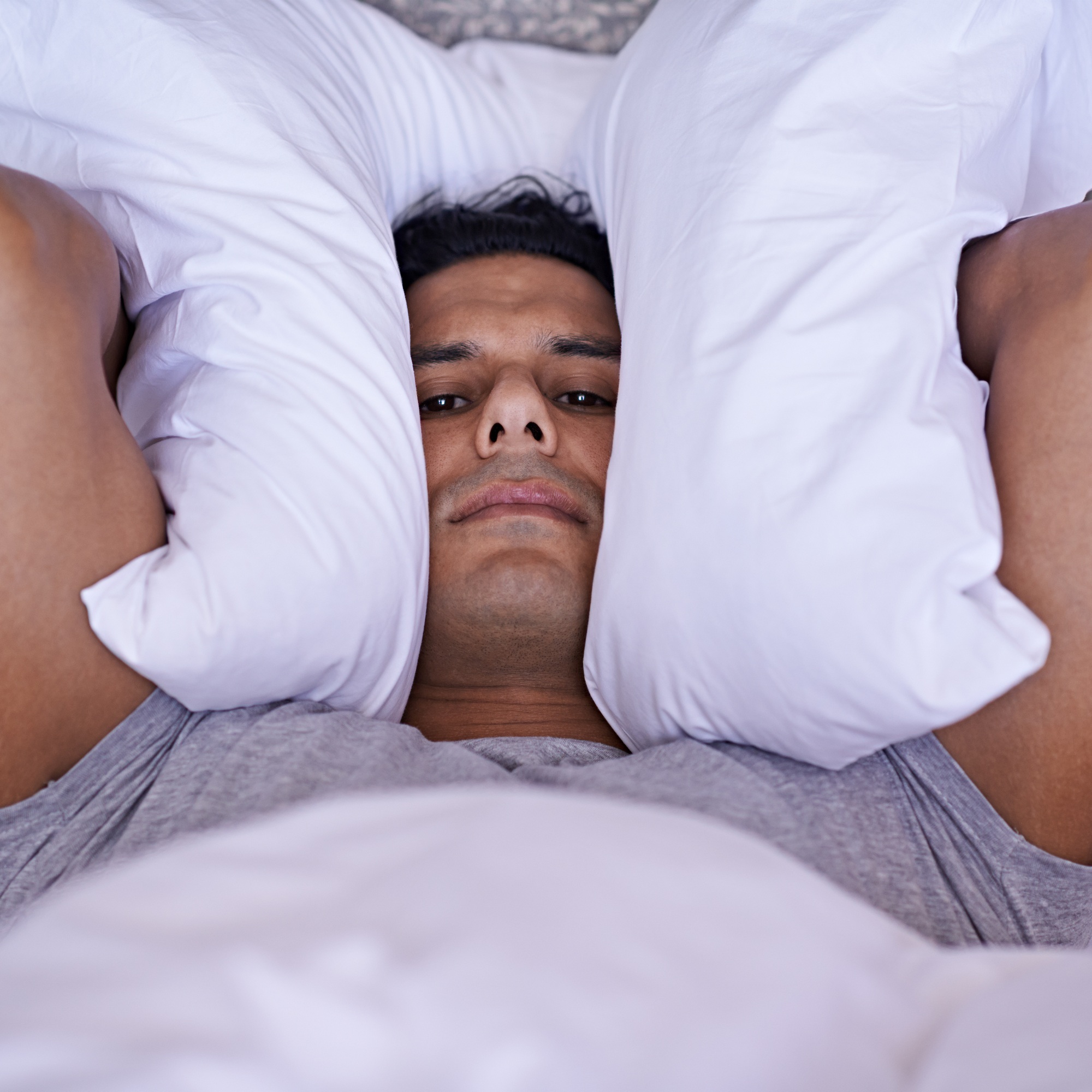 No-one can sleep with all this noise. A young man covering his ears with his pillow.
